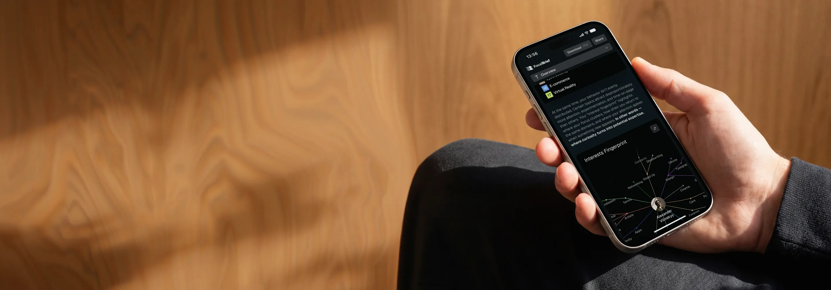 A hand holding a phone showing the FocalBrief interests fingerprint report, against a wooden wall